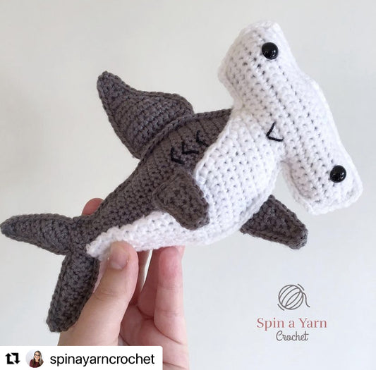 A small crochet gray and white smiling hammerhead shark is being held by a hand in front of a white background, the re-gram symbol is in the bottom left corner with the original creators social media handle; spinayarncrochet 