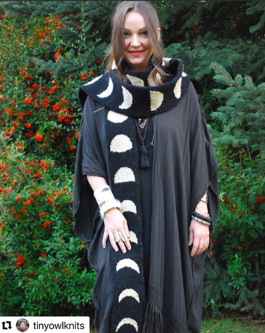A woman with brown hair stands outside in front of greenery modeling a black, long scarf with different phases of the moon in white pictured on it.