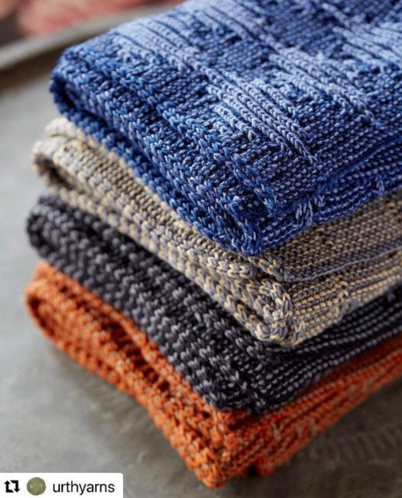 A stack of four different coloured Urth Synthesis Scarves sit on a gray table with a re-gram symbol in the bottom left corner with the original handle @urthyarns listed.