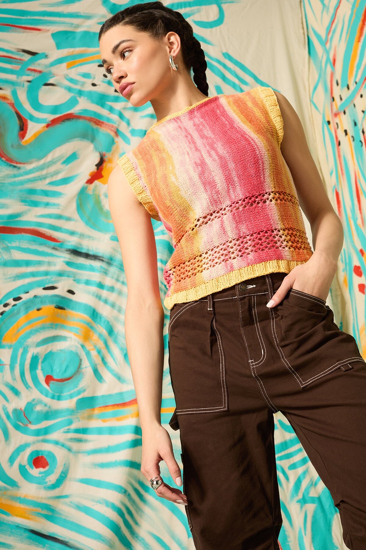 Woman wearing a colorful sleeveless top and brown pants against a vibrant, abstract wall.