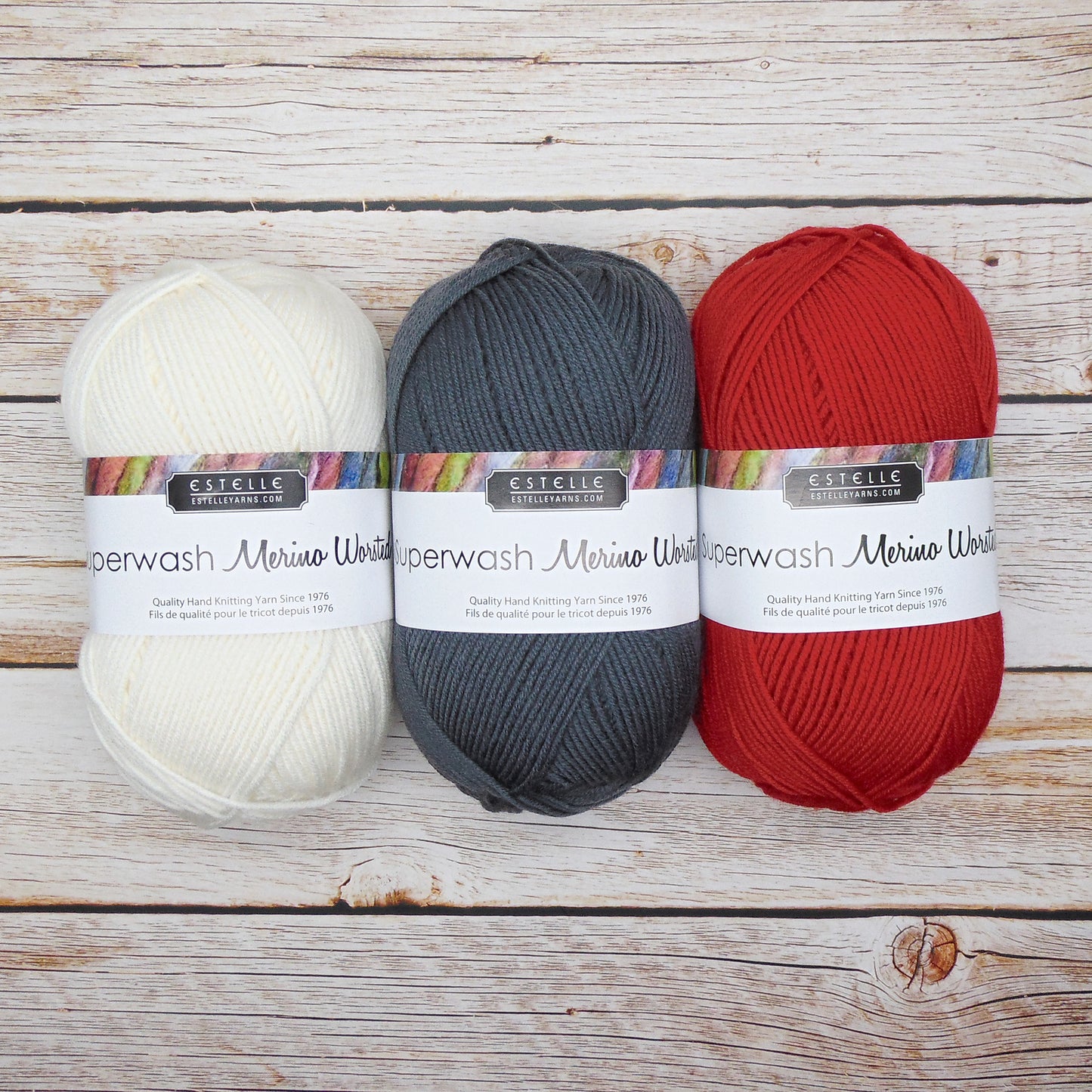 Three balls of Estelle Superwash Merino Wool yarn in white, gray, and red on a wooden surface.