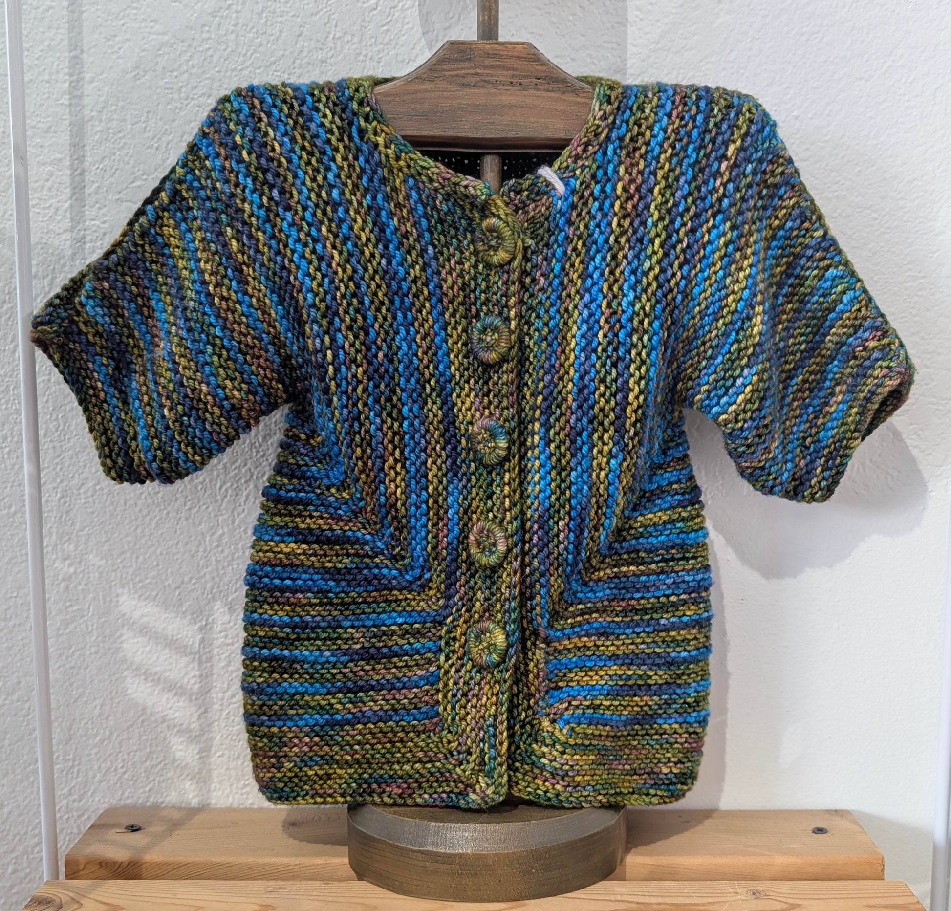 Knitted sweater on a mannequin against a white wall with wooden shelves below.